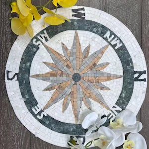 Può includere: Una rosa dei venti a mosaico rotonda con un motivo a stella nei toni neutri del grigio, beige e marrone. La bussola è circondata da una fascia verde scuro con lettere bianche che indicano le direzioni. Orchidee gialle e bianche sono disposte attorno alla bussola.