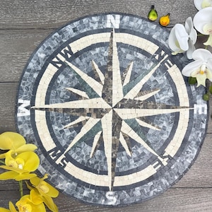 Peut inclure: Boussole en mosaïque ronde avec un schéma de couleurs gris, crème et bleu foncé. Le motif de la rose des vents comporte des indicateurs directionnels. Des orchidées jaunes et blanches et de petits oiseaux décoratifs sont disposés autour de la boussole.
