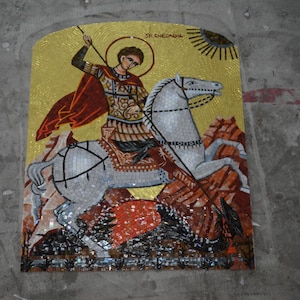 Puede incluir: Mosaico que representa una figura a caballo, sosteniendo una lanza, con un halo dorado y un diseño de sol. El mosaico presenta un diseño detallado con varios colores y texturas, montado sobre una superficie gris.