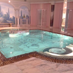 Puede incluir: Una piscina cubierta con un diseño floral de mosaico en el suelo. La piscina tiene una cubierta de madera y está rodeada de suelos de mármol y columnas. Se ve un mural de un paisaje.
