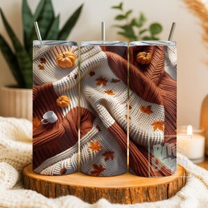 Op de afbeelding: Een cilindervormige beker met rietje voorzien van een print van een wit en roestkleurig gebreid truipatroon, verspreide oranje herfstbladeren, kleine oranje pompoenen en een piepklein wit kopje. Het ontwerp bedekt het gehele oppervlak.