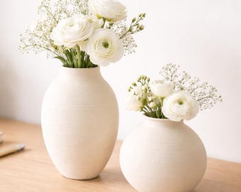 May include: Two off-white ceramic vases, one tall and one round, each filled with white flowers and greenery. The vases sit on a light wood surface, with a small potted plant and a notebook visible in the background.