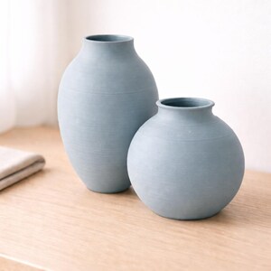 Pu&ograve; includere: Due vasi in ceramica azzurra di diverse dimensioni. Il vaso pi&ugrave; alto ha una forma cilindrica, mentre quello pi&ugrave; piccolo &egrave; rotondo. Entrambi i vasi hanno una superficie strutturata e poggiano su una superficie di legno.