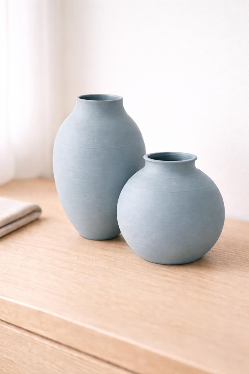 Pu&ograve; includere: Due vasi in ceramica azzurra di diverse dimensioni. Il vaso pi&ugrave; alto ha una forma cilindrica, mentre quello pi&ugrave; piccolo &egrave; rotondo. Entrambi i vasi hanno una superficie strutturata e poggiano su una superficie di legno.