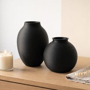 Pu&ograve; includere: Due vasi in ceramica nera opaca di diverse dimensioni sono su una superficie di legno. Una candela accesa in un barattolo di vetro &egrave; accanto ai vasi. Un tovagliolo di lino e spighe di grano secche sono anche sul tavolo.
