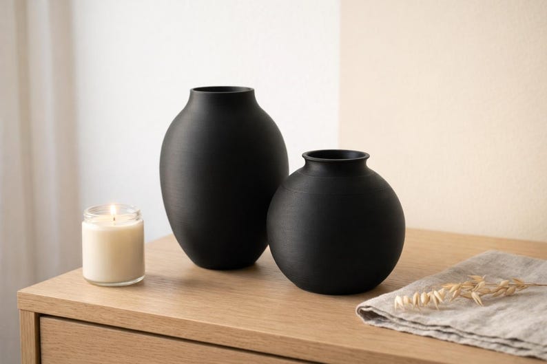 Pu&ograve; includere: Due vasi in ceramica nera opaca di diverse dimensioni sono su una superficie di legno. Una candela accesa in un barattolo di vetro &egrave; accanto ai vasi. Un tovagliolo di lino e spighe di grano secche sono anche sul tavolo.