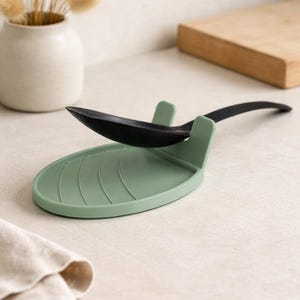 Può includere: Un poggia cucchiaio verde salvia con un cucchiaio nero. Il poggia cucchiaio ha una forma ovale curva con creste rialzate e due piccoli supporti verticali. Il cucchiaio è appoggiato sul poggia cucchiaio. Articoli per la cucina.