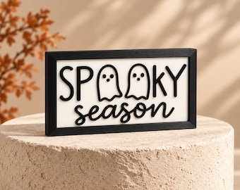 May include: A rectangular sign with a black frame and a white background. The sign features black raised lettering that reads "SPOOKY season," with two ghost shapes replacing the letter "O" in the word "SPOOKY." The design is clean and simple.