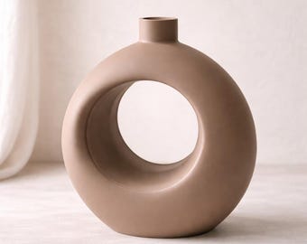 May include: A modern, taupe-colored ceramic vase with a unique circular design. The vase features a rounded body with a large, open circle in the center and a small cylindrical opening at the top. This decorative piece adds a contemporary touch.
