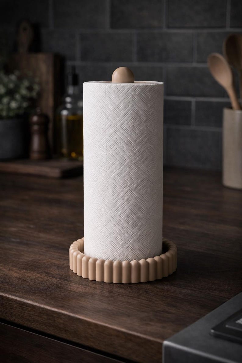 Puede incluir: Un soporte vertical para papel de cocina con una base circular estriada en color beige claro y un remate esf&eacute;rico a juego en la parte superior. El soporte contiene un rollo de papel blanco con un patr&oacute;n geom&eacute;trico en relieve.