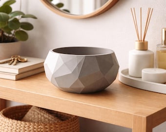 May include: A gray geometric bowl sits on a wooden table, alongside a plant, books, and decorative items. The bowl's faceted design adds a modern touch to the decor. A round mirror is visible in the background.