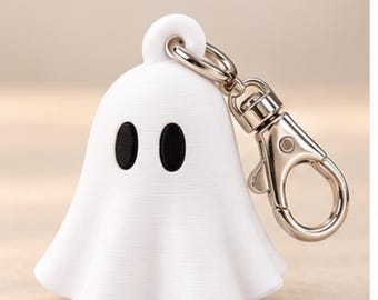May include: A white ghost-shaped keychain featuring two oval black eyes and a silver-toned metal lobster clasp attached to the top. The ghost figure has a smooth, rounded form with subtle horizontal texture lines across its surface.