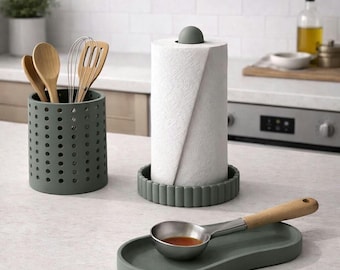 May include: A kitchen countertop scene featuring a gray paper towel holder, utensil holder, and spoon rest. The paper towel holder has a gray base and a gray knob. The utensil holder is cylindrical with multiple holes. A stainless steel spoon rests on a gray tray.