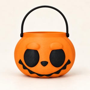 May include: A bright orange pumpkin-shaped bucket with a black handle. The bucket features a moulded face with large, round black eyes, a small triangular nose, and a wide, jagged black mouth. The surface has a visible 3D printed texture.
