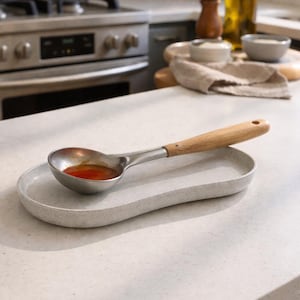 Puede incluir: Un cucharón de acero inoxidable con mango de madera descansa sobre un reposacucharas de cerámica gris moteada. El cucharón contiene un líquido rojo. La encimera de la cocina y el fondo son visibles.