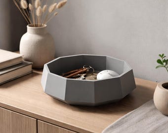 May include: A gray, geometric-shaped catch-all tray holding keys and a small white object. The tray sits on a wooden surface, with a vase of dried flowers and a small plant in the background. A stylish storage solution.