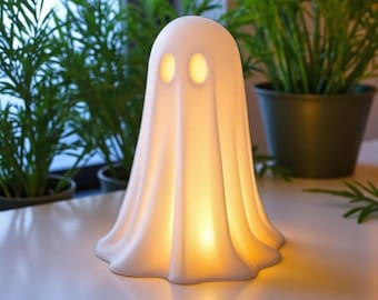 May include: A white, ghost-shaped lamp with a smooth, matte finish. The lamp features two oval-shaped cutouts for eyes and emits a warm, soft yellow glow from within, illuminating the structure and casting a gentle light on the surface below.