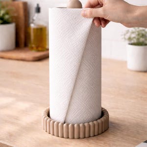 Puede incluir: Un rollo de papel de cocina blanco sobre un soporte beige con un borde decorativo y acanalado. El rollo est&aacute; rematado con un peque&ntilde;o pomo beige redondo. El soporte est&aacute; sobre una encimera de madera clara en una cocina.