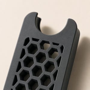 May include: A dark gray, rectangular phone accessory with a honeycomb pattern and a curved top. The accessory has a series of hexagonal cutouts, creating a textured surface. The design is modern and minimalist.