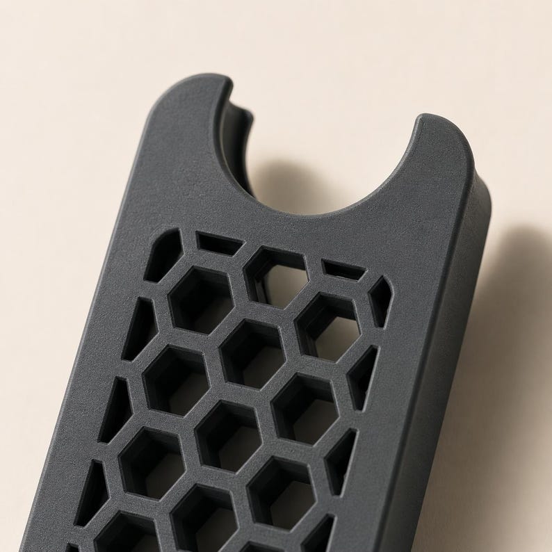 May include: A dark gray, rectangular phone accessory with a honeycomb pattern and a curved top. The accessory has a series of hexagonal cutouts, creating a textured surface. The design is modern and minimalist.