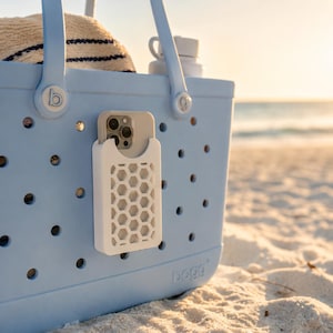 May include: A light blue tote bag with a white phone holder attached. The phone holder has a honeycomb design. A phone is visible in the holder. The bag is on a sandy beach with a hat and water bottle inside.