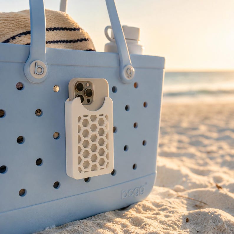 May include: A light blue tote bag with a white phone holder attached. The phone holder has a honeycomb design. A phone is visible in the holder. The bag is on a sandy beach with a hat and water bottle inside.