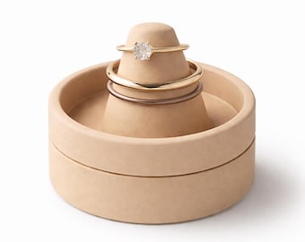 May include: A beige ring holder with a conical top and a circular base. Three rings are displayed on the holder: a gold band with a diamond, a gold band, and a silver band. The holder is made of a smooth, matte material.