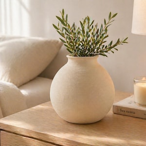 May include: A beige, round ceramic vase with a textured surface, filled with green leafy branches. The vase sits on a wooden nightstand next to a lit candle in a glass holder and a book titled "Bea moma".