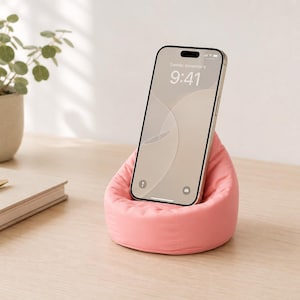 May include: A pink beanbag-style phone stand supporting a smartphone. The phone screen shows 9:41. A notebook, pen, and a potted plant are on a wooden surface.