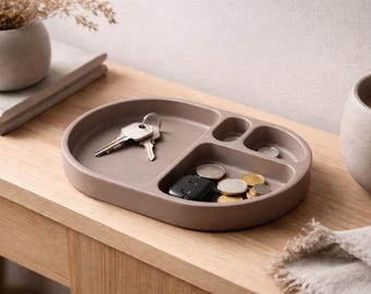 May include: A taupe-colored oval tray with multiple compartments, holding keys, coins, and a car key fob. The tray sits on a wooden surface, with a small vase and a mug visible in the background. The tray is designed for organizing small items.