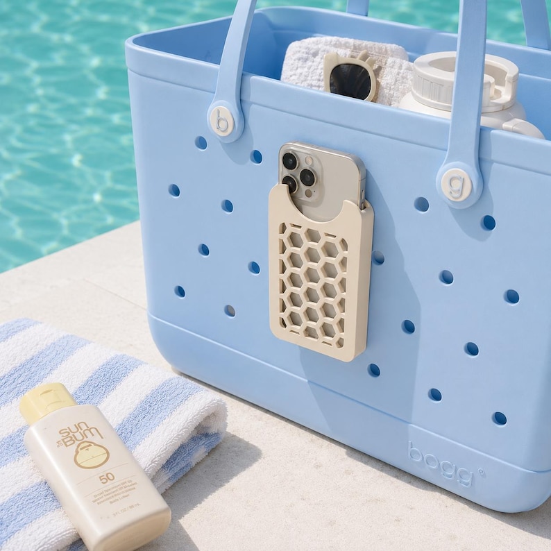 May include: A light blue tote bag with a beige honeycomb phone holder attached. The bag contains a white towel, sunglasses, and a water bottle. A bottle of Sun Bum sunscreen and a blue and white striped towel are in the foreground.