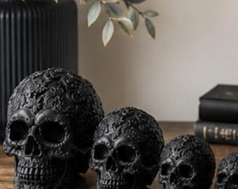 May include: A set of four matte black skulls of varying sizes, each featuring intricate, raised floral and scrollwork carvings across the entire surface. The skulls are arranged in a row from largest to smallest, displaying detailed eye sockets and teeth.