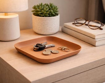 May include: A square, terracotta-colored tray holds keys, coins, and rings on a wooden nightstand. A small succulent plant in a speckled white pot and a lamp with a beige shade are also on the nightstand. A pair of eyeglasses rests on a stack of books.