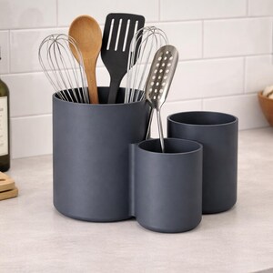 Puede incluir: Un juego de tres recipientes grises oscuros para utensilios de cocina sobre una encimera. Los recipientes contienen una cuchara de madera, una espátula negra, batidores y una cuchara ranurada. Una botella de aceite de oliva y una pequeña planta están en el fondo.