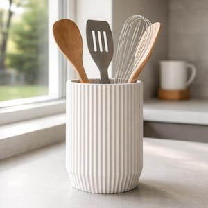 Puede incluir: Un recipiente blanco de cerámica acanalada para utensilios que contiene una cuchara de madera, una espátula gris, un batidor y otra cuchara de madera. El recipiente está sobre una encimera gris clara cerca de una ventana. Se ve una taza blanca en el fondo.
