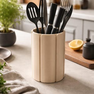 Puede incluir: Un soporte beige para utensilios con utensilios de cocina negros. El soporte es cilíndrico con crestas verticales y redondeadas. Los utensilios incluyen una cuchara ranurada, espátula, cuchillo y tenedores. Un limón y un pequeño recipiente negro.