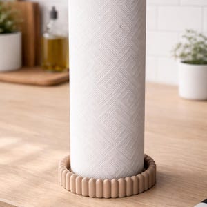 May include: A white paper towel roll with a geometric pattern sits on a beige, scalloped base. A small, round wooden knob tops the roll. The kitchen item is on a light wood surface.