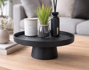 May include: A black, round, pedestal tray holds a small potted succulent, a candle labeled "Mindful #21", and a black diffuser bottle with reeds. The tray sits on a light wood coffee table. A vase of white flowers and books are also on the table.