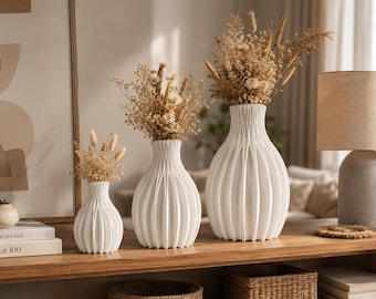May include: Three white ceramic vases of varying heights, each featuring a vertical ribbed texture and a narrow neck. Each vase contains a dried floral arrangement with beige and tan grasses, stems, and small flowers, creating a cohesive set.
