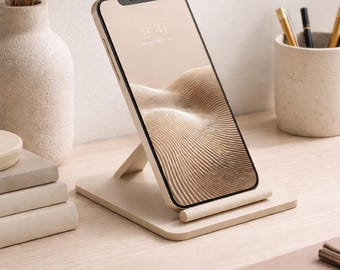 May include: A beige smartphone is displayed on a matching stand, showcasing a beige and brown abstract design on the screen. The phone is angled slightly, revealing its black frame. The stand is on a light wooden surface, with a vase and stationery nearby.