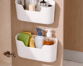 May include: Two white plastic storage baskets mounted on a wooden cabinet door. The top basket holds kitchen tools and spices. The bottom basket contains cleaning supplies, including a spray bottle and soap dispenser. The cabinet door is open.