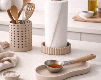 May include: A kitchen countertop scene featuring a beige utensil holder with wooden spoons and a whisk, a paper towel holder, and a spoon rest with a metal spoon. A small bowl of salt and a bottle of oil are also visible.