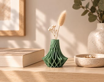 May include: A small, matte sage green vase with a geometric, lattice-like open structure. The vase has a wide, angular base that tapers upward to a narrow circular opening, containing a single dried beige grass stem and a small sprig of white flowers.