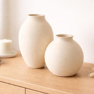 Pu&ograve; includere: Due vasi in ceramica bianco sporco di diverse dimensioni. Il vaso pi&ugrave; alto ha una forma ovale allungata, mentre quello pi&ugrave; piccolo &egrave; rotondo. Entrambi hanno una superficie strutturata e un design semplice e minimalista.