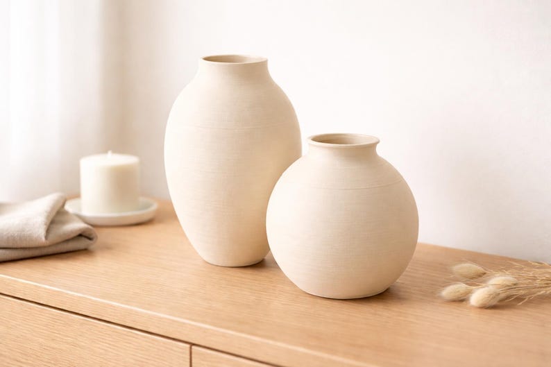Pu&ograve; includere: Due vasi in ceramica bianco sporco di diverse dimensioni. Il vaso pi&ugrave; alto ha una forma ovale allungata, mentre quello pi&ugrave; piccolo &egrave; rotondo. Entrambi hanno una superficie strutturata e un design semplice e minimalista.