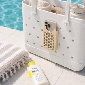 May include: A white tote bag with a beige phone holder attached, holding a phone. The bag has a handle and is near a striped towel and a bottle of sunscreen. The sunscreen bottle has yellow and blue accents and the text "PLAY".