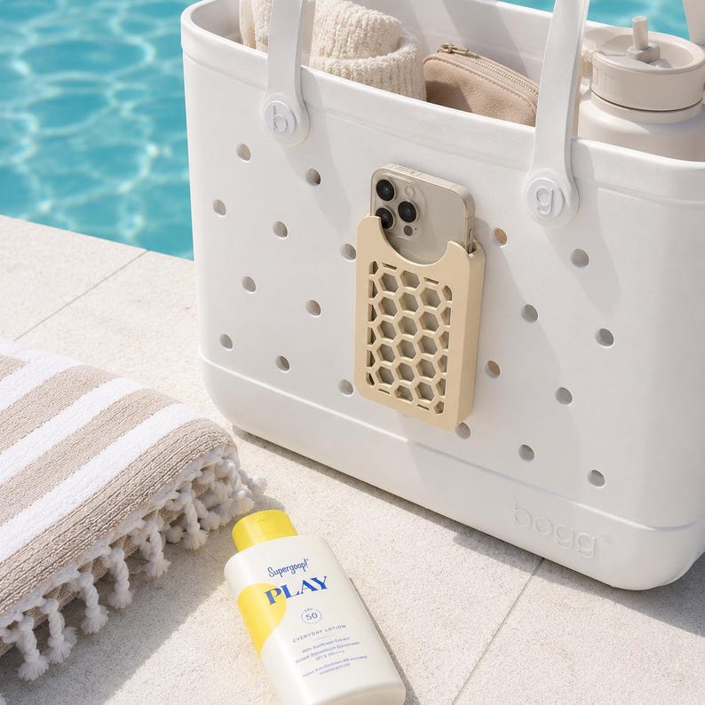 May include: A white tote bag with a beige phone holder attached, holding a phone. The bag has a handle and is near a striped towel and a bottle of sunscreen. The sunscreen bottle has yellow and blue accents and the text "PLAY".