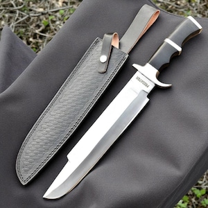 May include: A large hunting knife with a polished steel blade and a black handle, accompanied by a black leather sheath. The blade is marked with the word "PREDATOR". The sheath has a textured pattern and a leather strap.