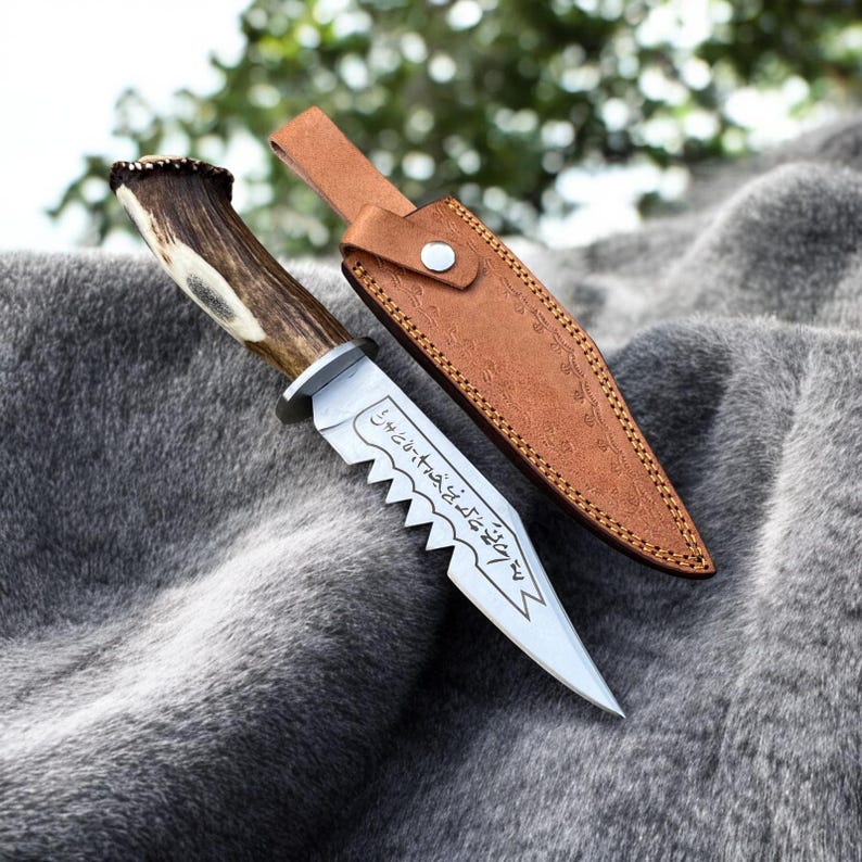 May include: A hunting knife with a serrated blade and a handle made from antler. The knife is accompanied by a brown leather sheath. The blade has etched text. The knife is displayed on a gray, furry surface.