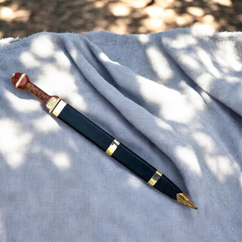 K&ouml;nnte beinhalten: Ein r&ouml;misches Gladius-Schwert mit schwarzer Scheide und goldfarbenen Akzenten. Der Griff ist braun mit einem roten Knauf. Das Schwert liegt auf einem grauen Tuch, mit Sonnenlicht.